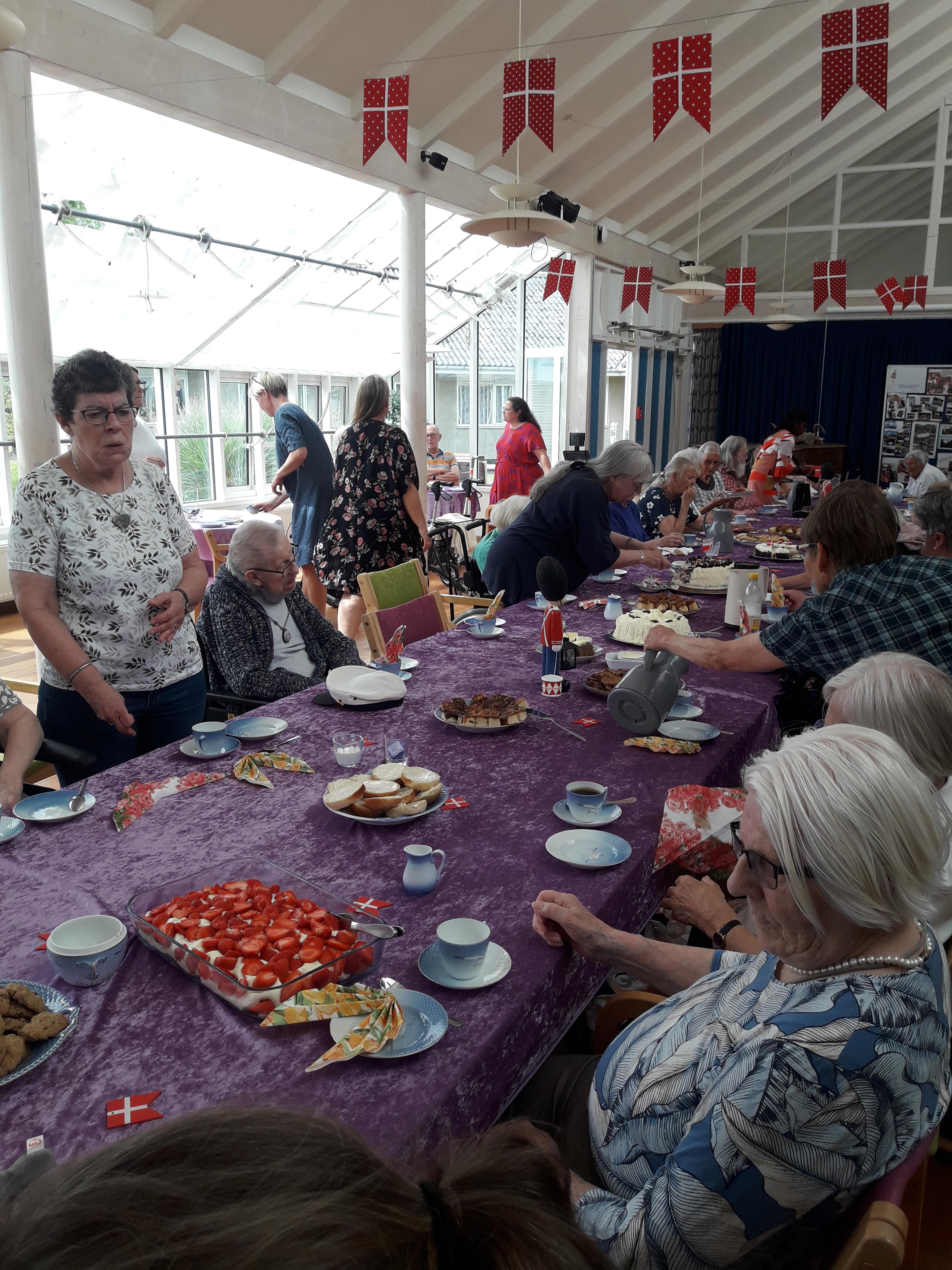 Beboere, frivillige og personale sidder ved pyntet bord med flag og kager Beboere, frivillige og personale sidder ved pyntet bord med flag og kager