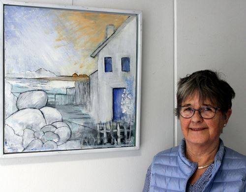 Månedens kunstner Inger Kruse ved et af sine udstillede malier af hus, vand og bjerg.