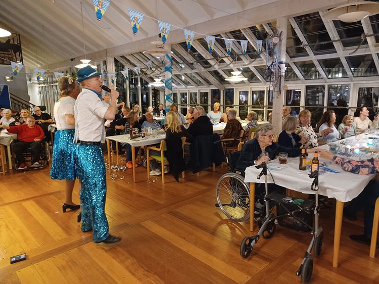 Beboere, pårørende, personale sidder ved borde og spiser. Der er pyntet til oktoberfest og underholdning af Stine og Martin. Beboere, pårørende, personale sidder ved borde og spiser. Der er pyntet til oktoberfest og underholdning af Stine og Martin.